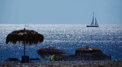 a sail boat in the ocean with a beach with umbrellas at Mojito Beach Rooms in Lachania