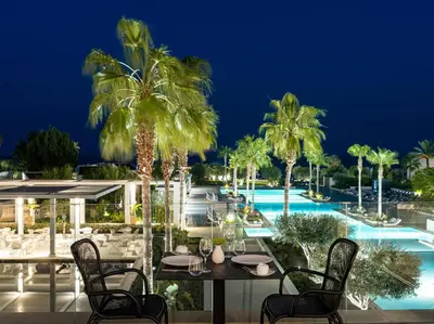 a restaurant with palm trees and a pool at night at Grecotel LUXME Dama Dama in Faliraki