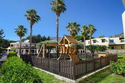 a playground behind a fence with palm trees at Pefkos Village Resort in Pefki Rhodes