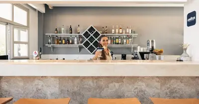 a woman standing behind a bar holding a cell phone at Pedi Beach Hotel in Symi