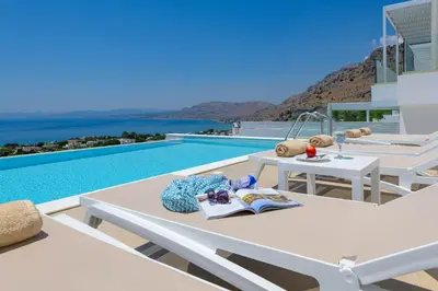 a pool with white chairs and a table with a book at Villa Dione in Pefkos, Lindos area in Líndos