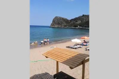 a wooden bench on a beach with people in the water at stegna on sea apt in Archangelos